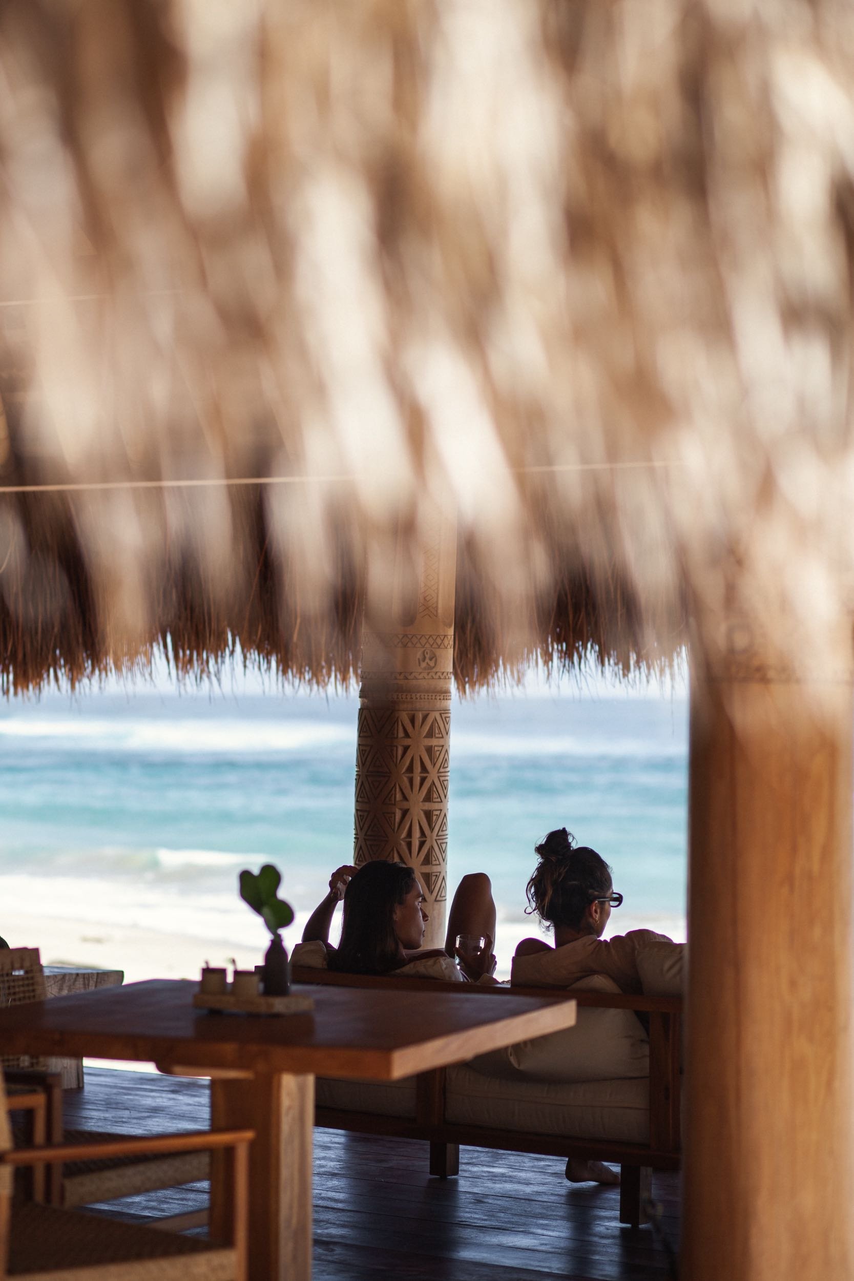 People sitting under a thatched roof facing the ocean at a beachside restaurant.