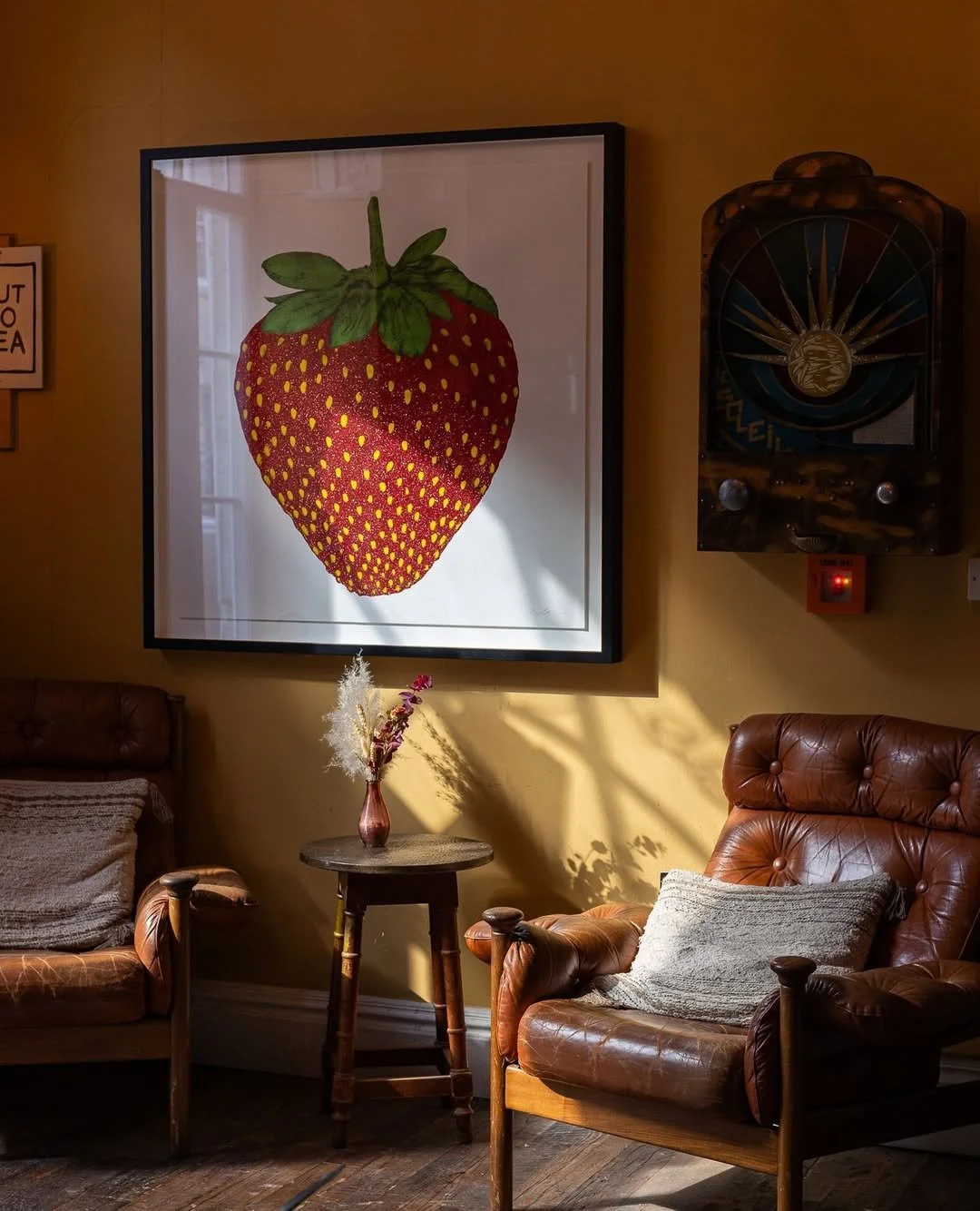 Interior of a cozy room with leather armchairs, a small wooden table with a vase of dried flowers, and large framed artwork of a strawberry on the wall, with sunlight casting shadows.