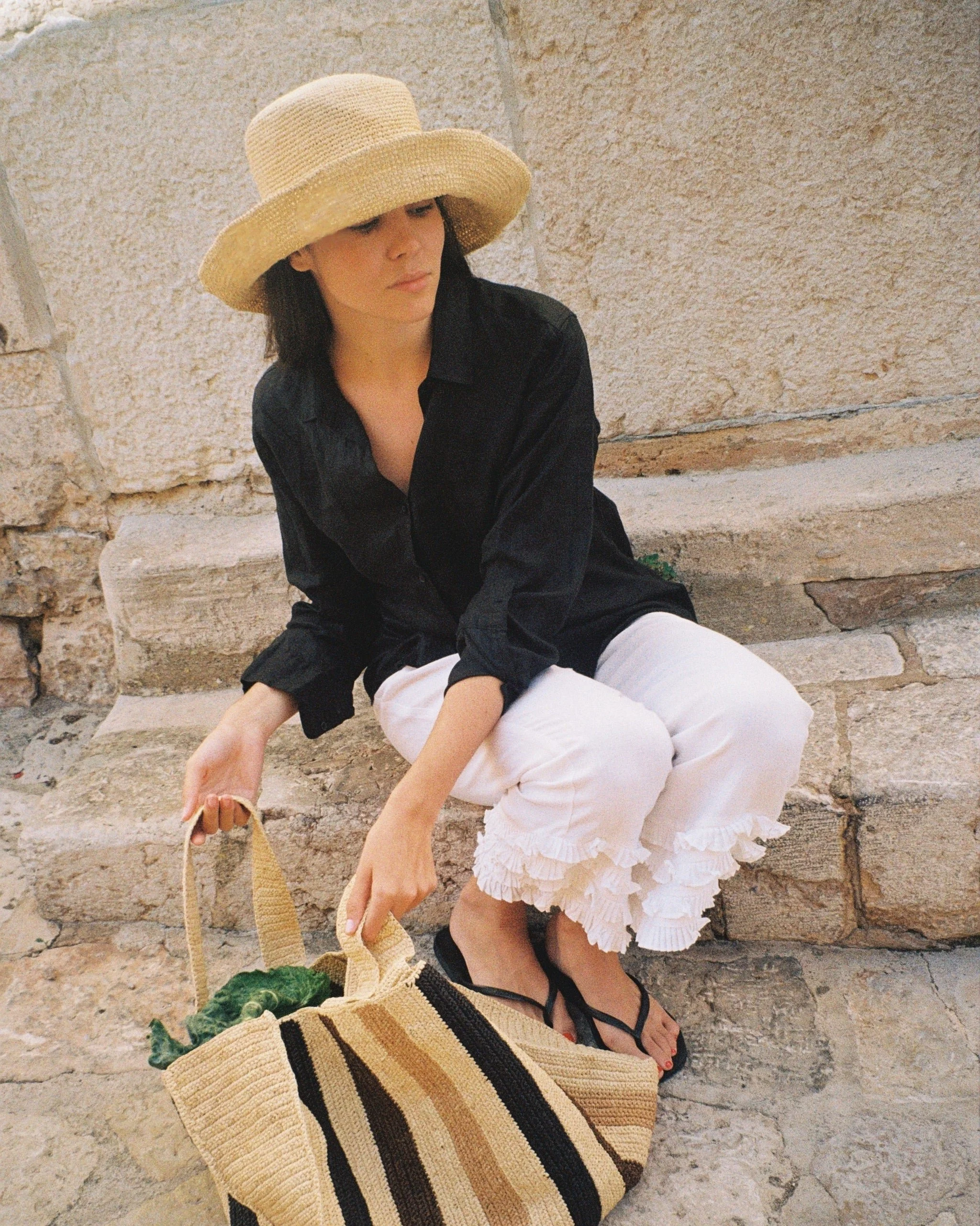 A woman wearing a wide-brimmed straw hat, black top, and white ruffled shorts sitting on stone steps beside a beige stone wall, holding a large straw bag.