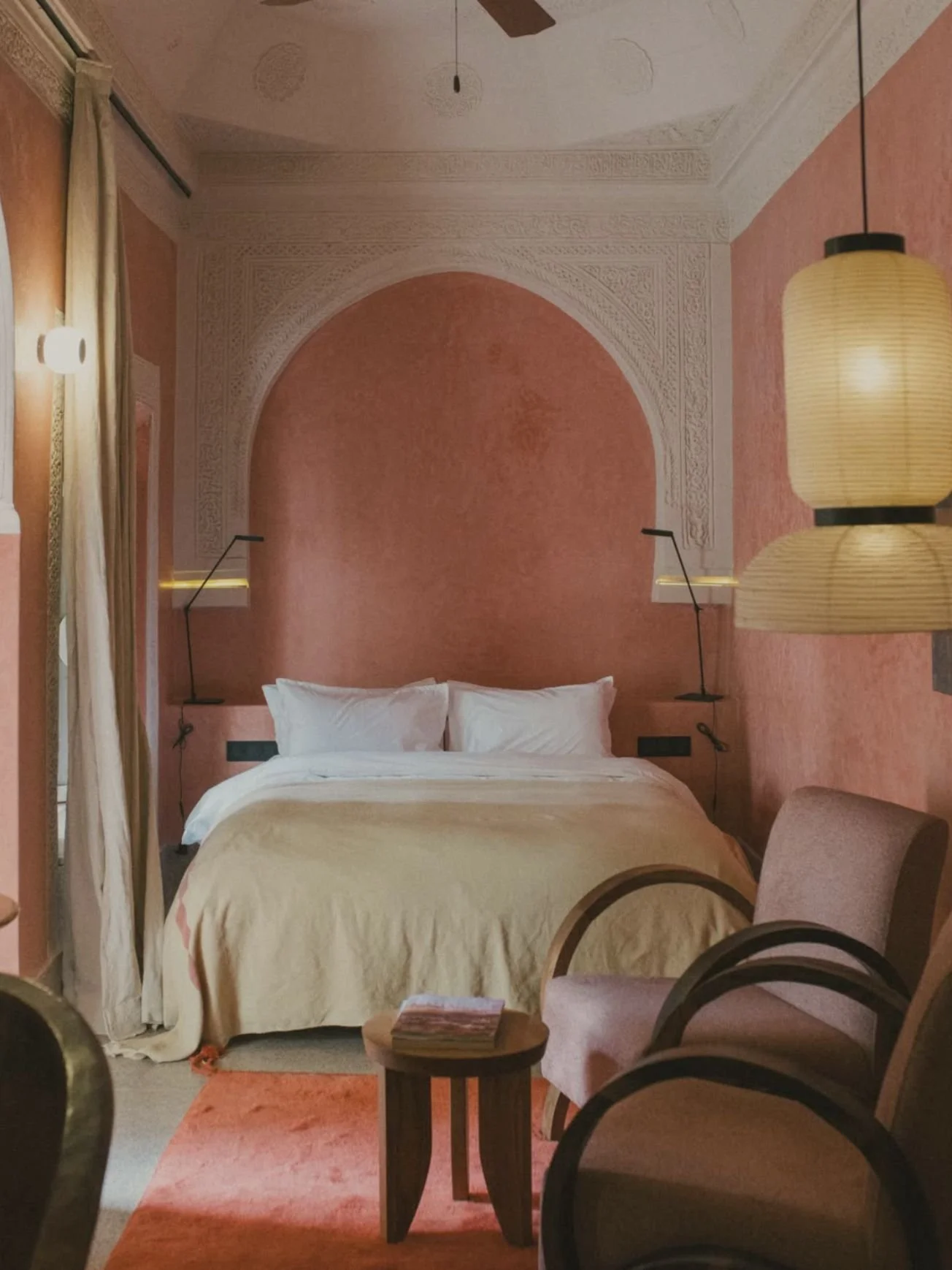 A cozy hotel bedroom with pink textured walls, a white bed with plush pillows, a beige blanket, and vintage-style hanging lamps on each side of the bed. There is a small wooden table in front of the bed with a few magazines, and a pink carpet on the floor.