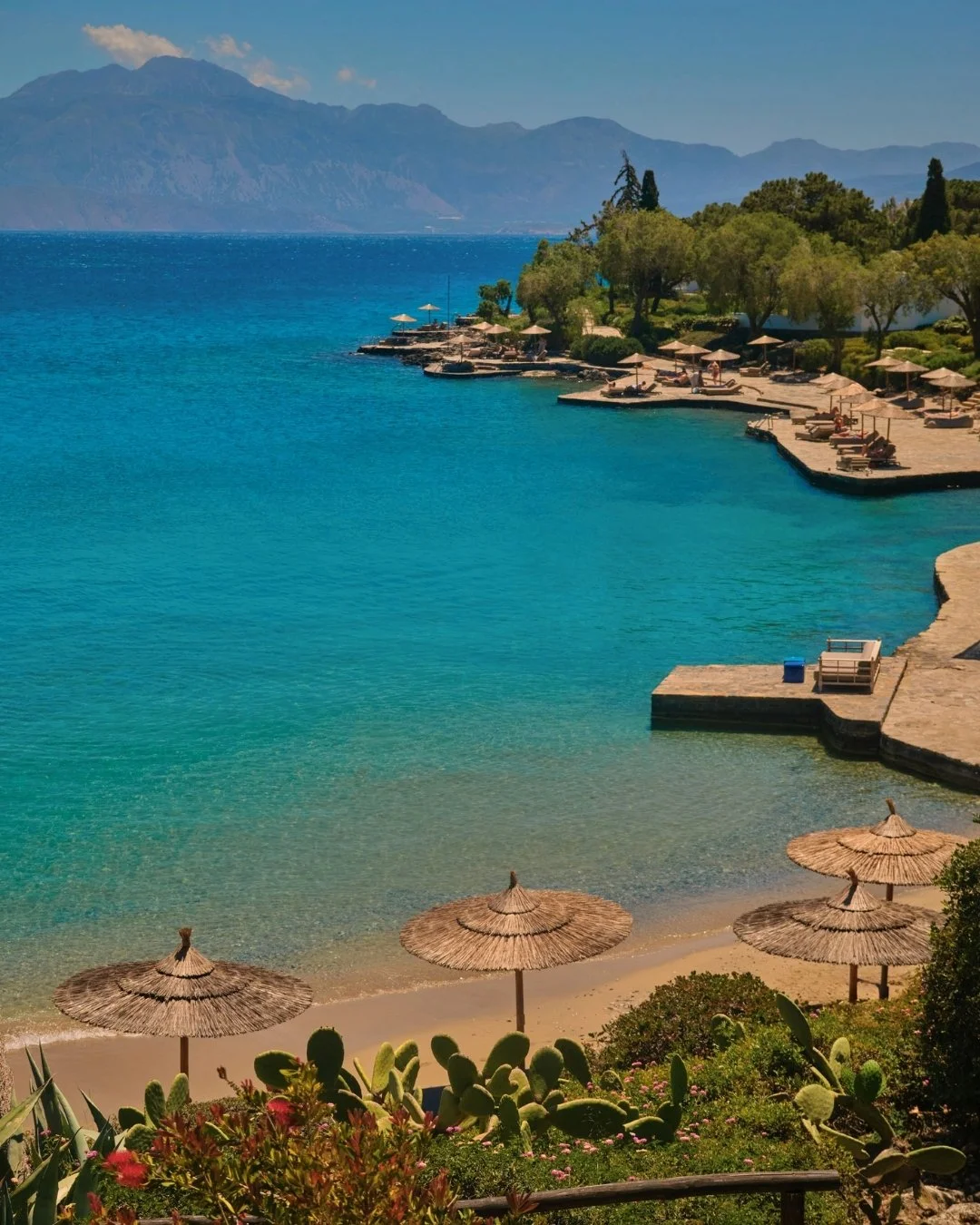 Beach with straw umbrellas, clear blue water, trees, lounge chairs, and mountains in the background.