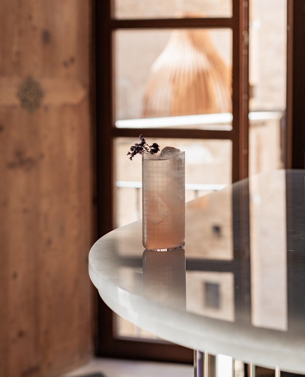 A tall glass with a grid pattern, filled with a pink-colored beverage, ice, and garnished with a small branch of purple flowers, sitting on a white marble table near a wooden window.