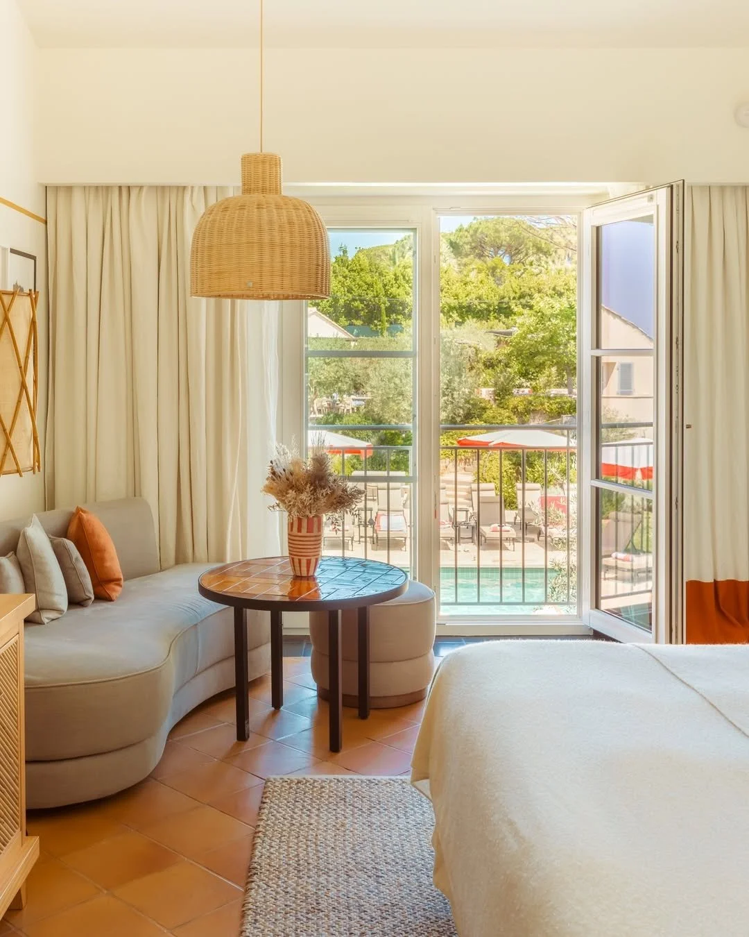 A cozy hotel room with a sliding glass door leading to a balcony with outdoor chairs and umbrellas, a beige curved sofa with pillows, a wooden round coffee table with a vase of dried flowers, terracotta tiled floor, and cream-colored curtains.
