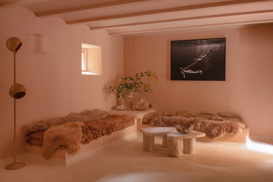 Cozy corner with two plush fur-covered benches, a small round table with a rock on it, a potted plant, a window, a wall art piece, and a standing lamp, all in soft beige tones.