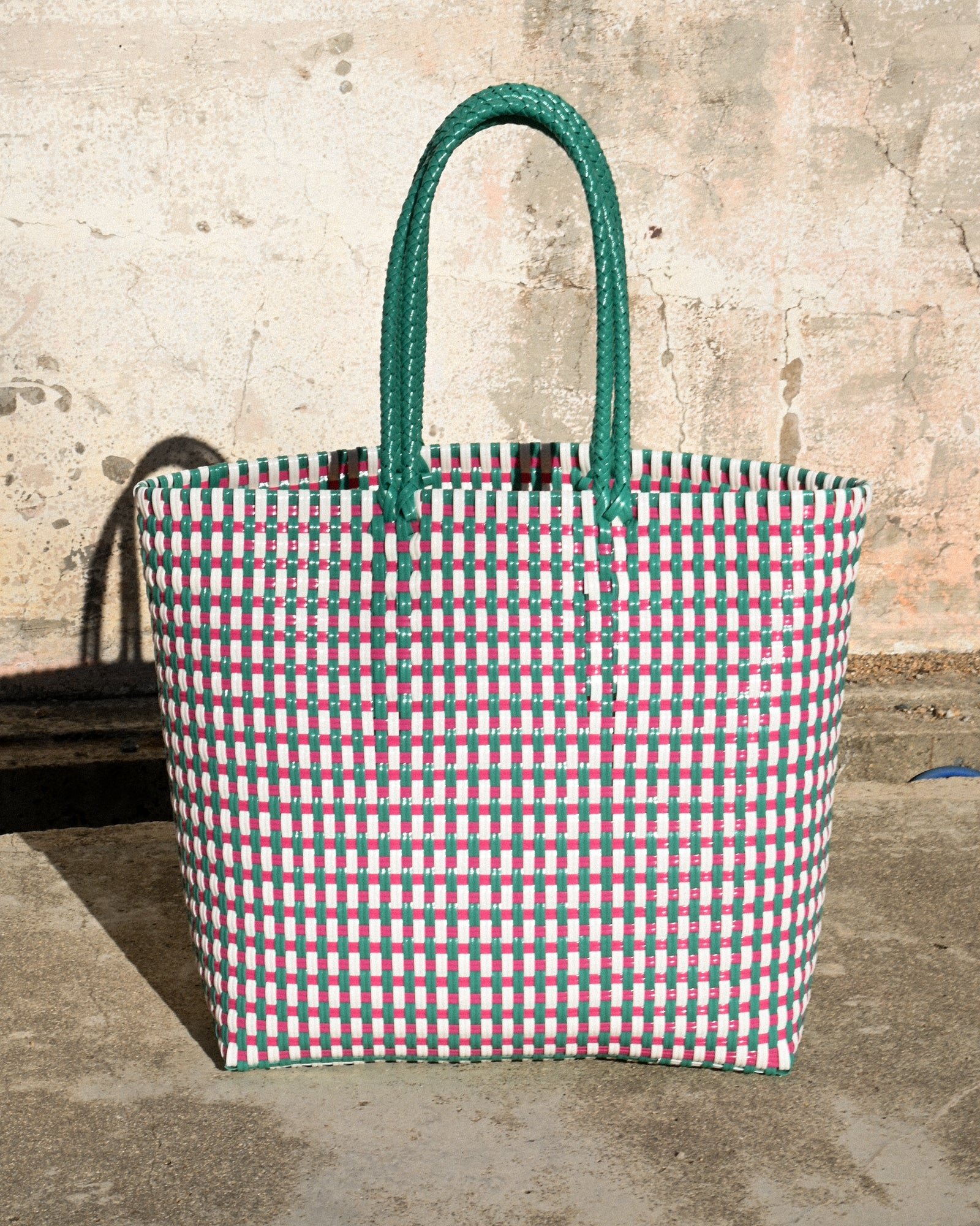 A woven fabric tote bag with a red, green, and white checkered pattern. It has green handles made of braided material and hangs against a textured, weathered beige wall.