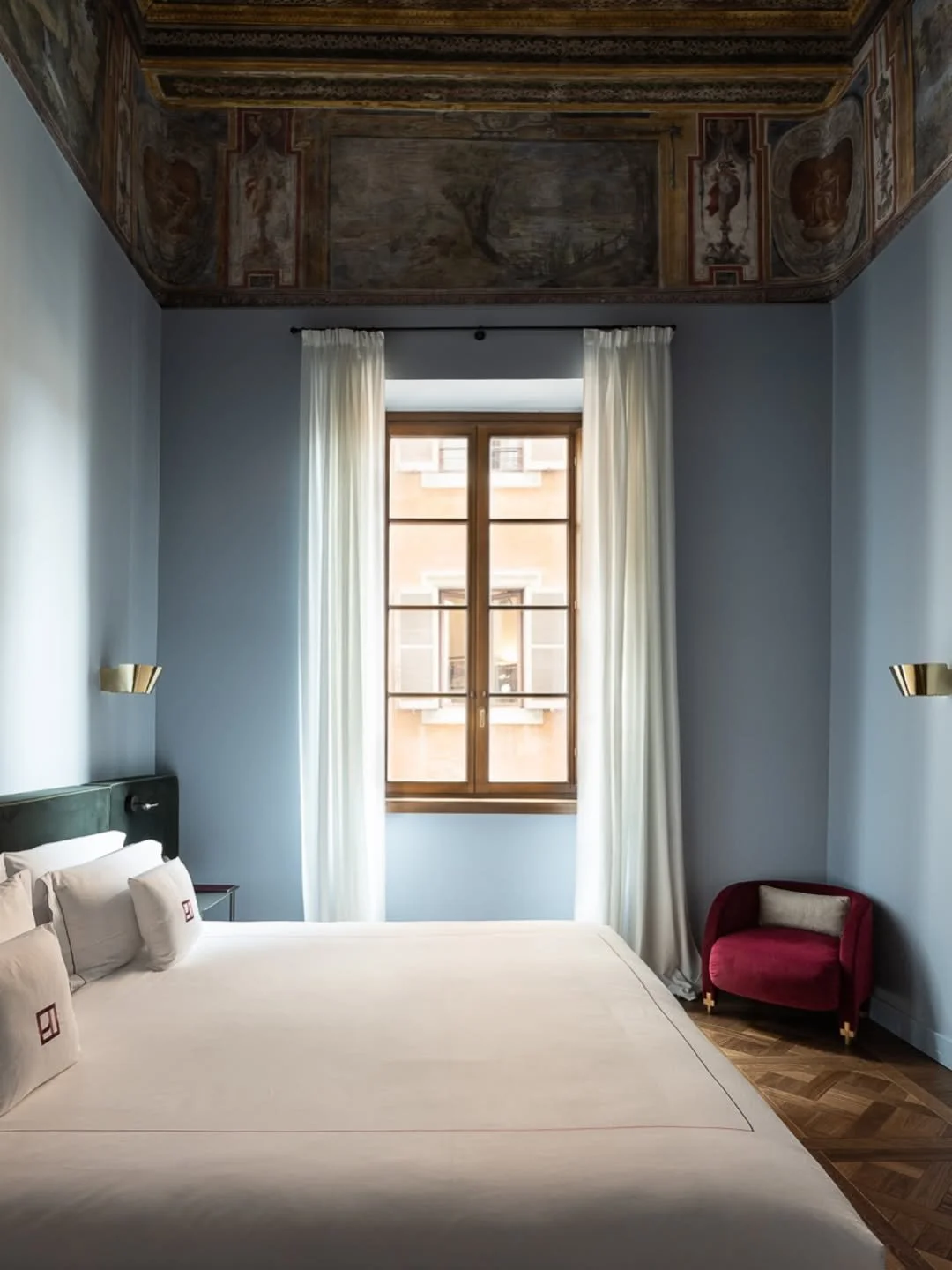 A bedroom with high ceilings and a large window with white curtains, a bed with white linens and pillows, a red armchair with a beige pillow, and ornate frescoes on the ceiling.