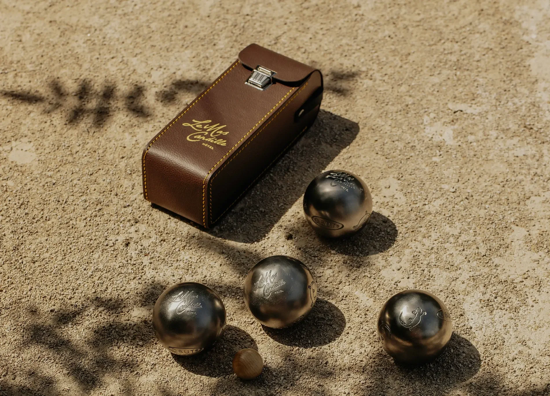 A vintage-style brown leather box with yellow stitching and gold lettering reading 'Le Mas Castaill' on a sandy outdoor surface. Several metallic petanque balls, engraved with designs and the words 'Le Mas Castaill' and 'NOTE,' are scattered nearby in the sand.