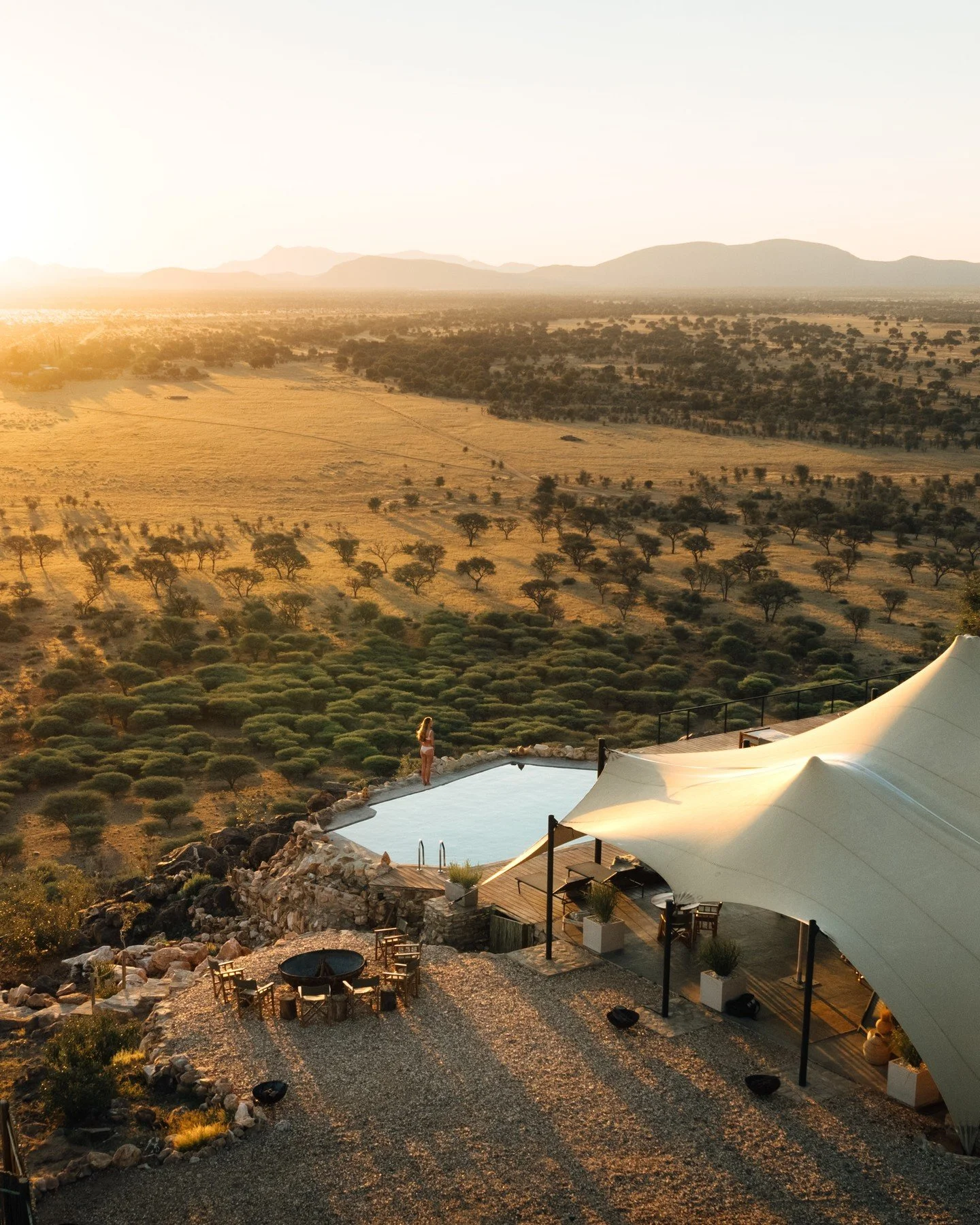 Sunset view of a vast desert landscape with scattered trees and distant mountains, featuring a pool and a white canopy in the foreground.