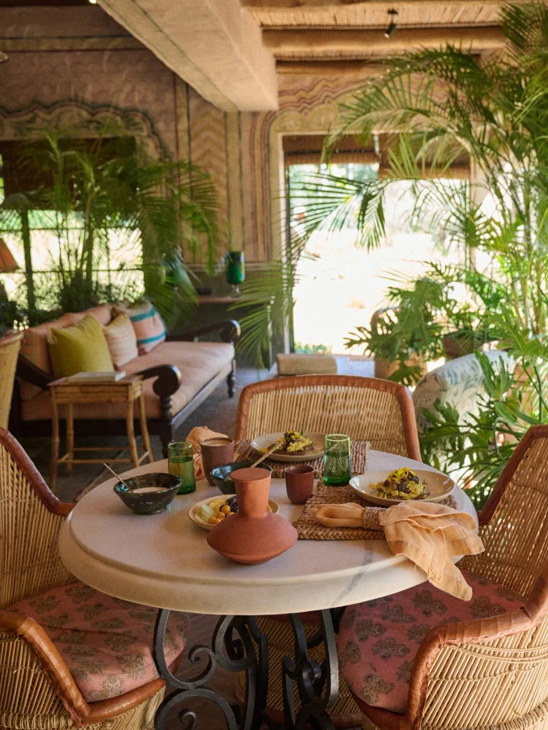 Indoor dining area with a round table set for a meal, surrounded by wicker chairs, lush green plants, a sofa with colorful cushions, and large windows letting in natural light.