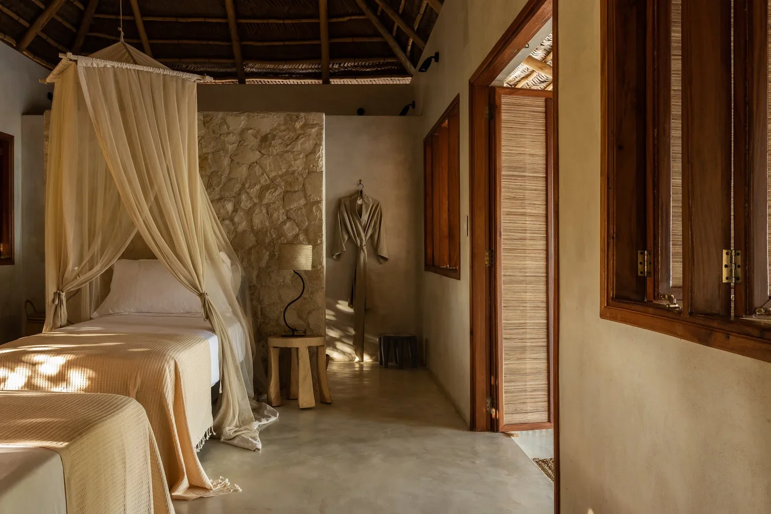 A bedroom with a bed draped with a canopy, light-colored bedding, a rustic stone wall, wooden windows, and a robe hanging on the wall.