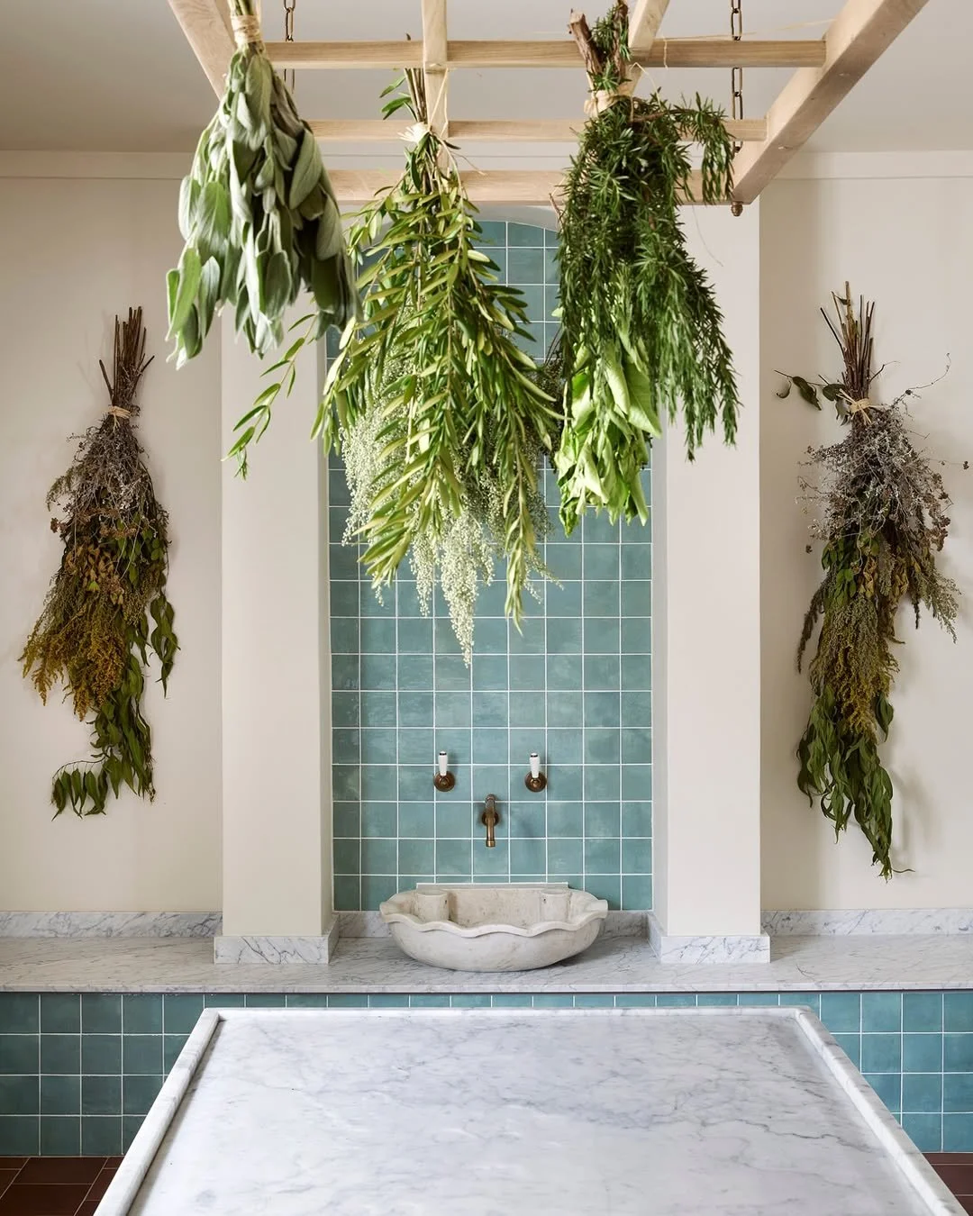 Hanging bundles of fresh and dried herbs above a small marble basin with blue-green tile background and white walls.
