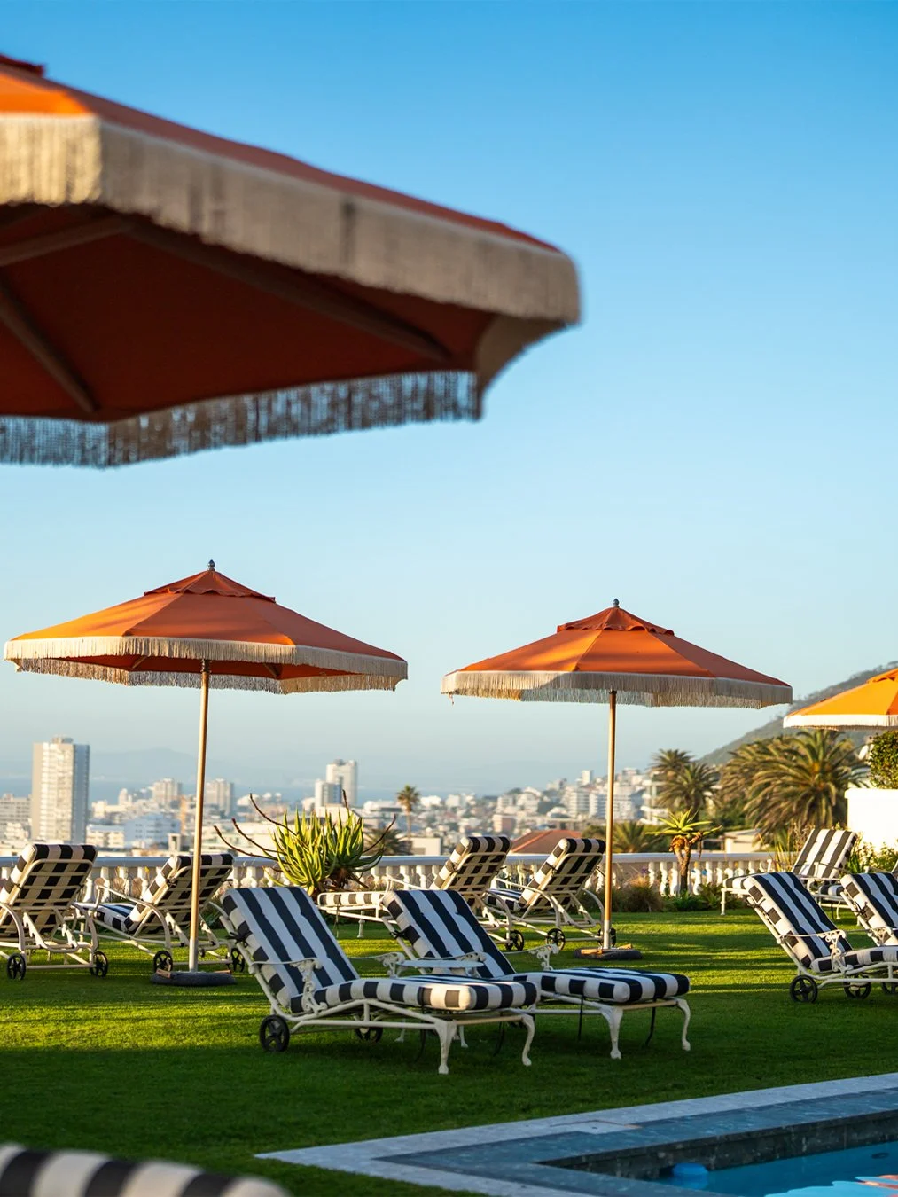 Poolside with black and white striped lounge chairs, large orange umbrellas, and a city skyline in the background against a clear blue sky.
