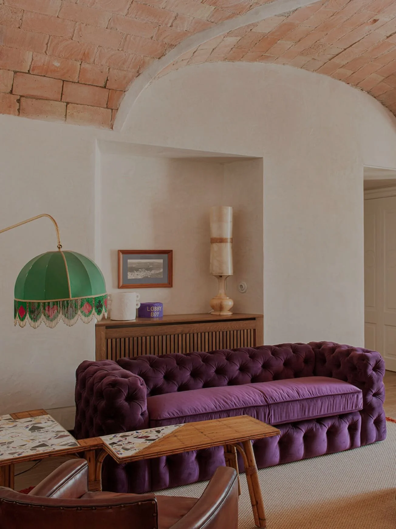 Interior living space with a purple velvet tufted sofa, wooden table, green hanging lamp with decorative fringe, beige wall with built-in shelf, framed picture, tall textured lamp, and brick arch ceiling.
