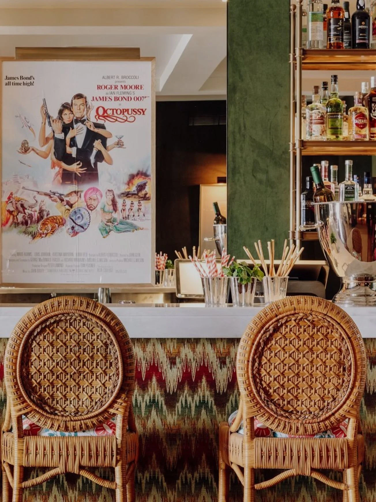 Interior of a bar or restaurant with two wicker chairs in the foreground, a colorful patterned cloth below, and a bar counter with glassware, straws, and a large photo poster of James Bond from the movie Octopussy on the wall. Shelves with various bottles of alcohol are in the background.