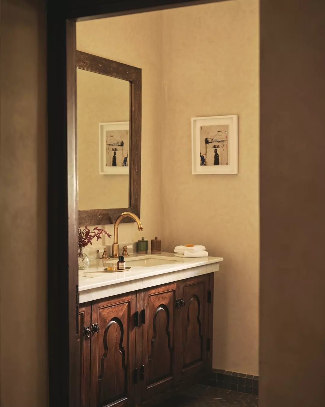 A bathroom vanity with a marble top, dark wooden cabinet doors, a gold faucet, and a round mirror, with framed artwork on the beige wall and folded towels on the right side.