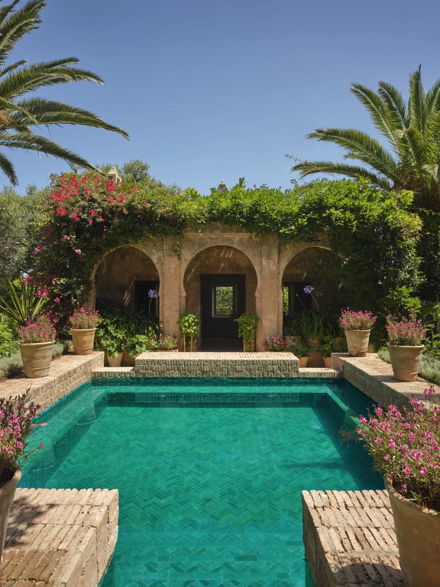 View of a swimming pool in a lush garden with a stone building, pink and purple flowers, and palm trees under a clear blue sky.