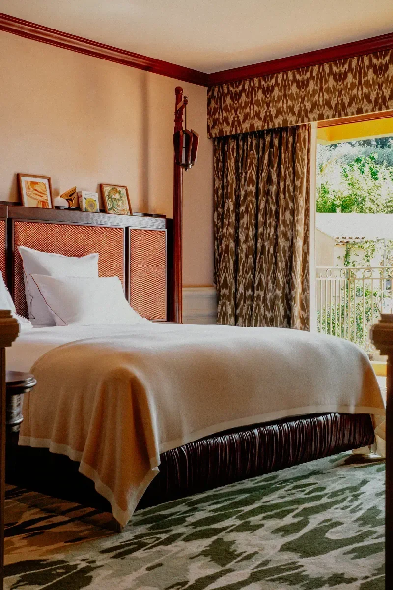 Interior of a hotel room with a neatly made bed, decorative curtains, and a balcony door showing greenery outside.