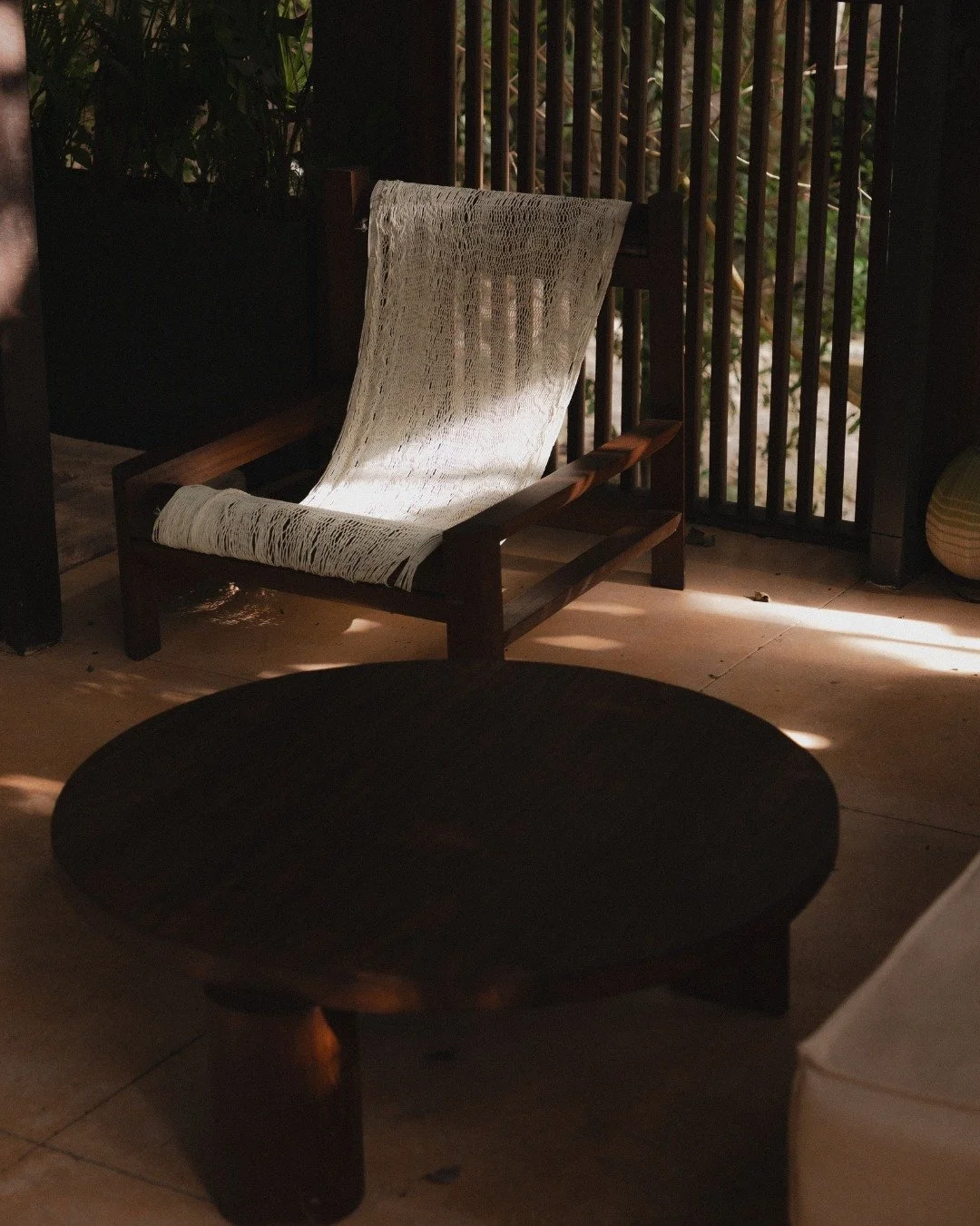 A cozy outdoor seating area with a wooden chair draped with a white blanket, sunlight streaming through a slatted wooden fence, and a dark round table in the foreground.