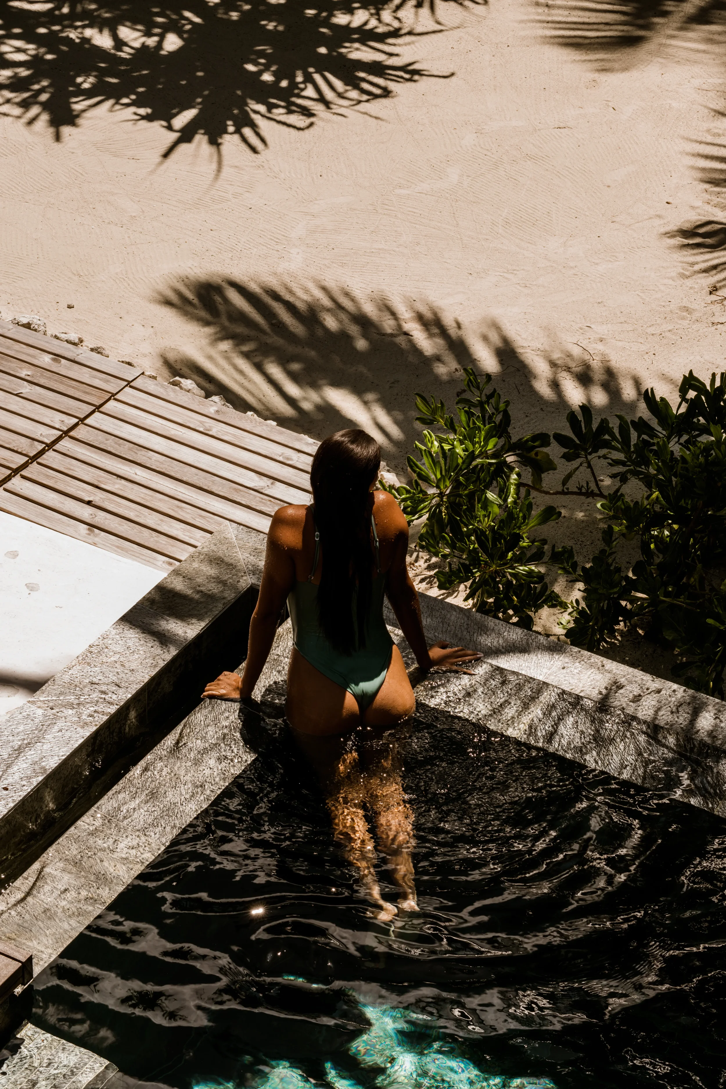 A woman with long dark hair in a swimsuit sits on the edge of a small pool, with her back facing the camera. Shadows of palm trees are cast onto the sandy area nearby, and nearby green foliage is visible.