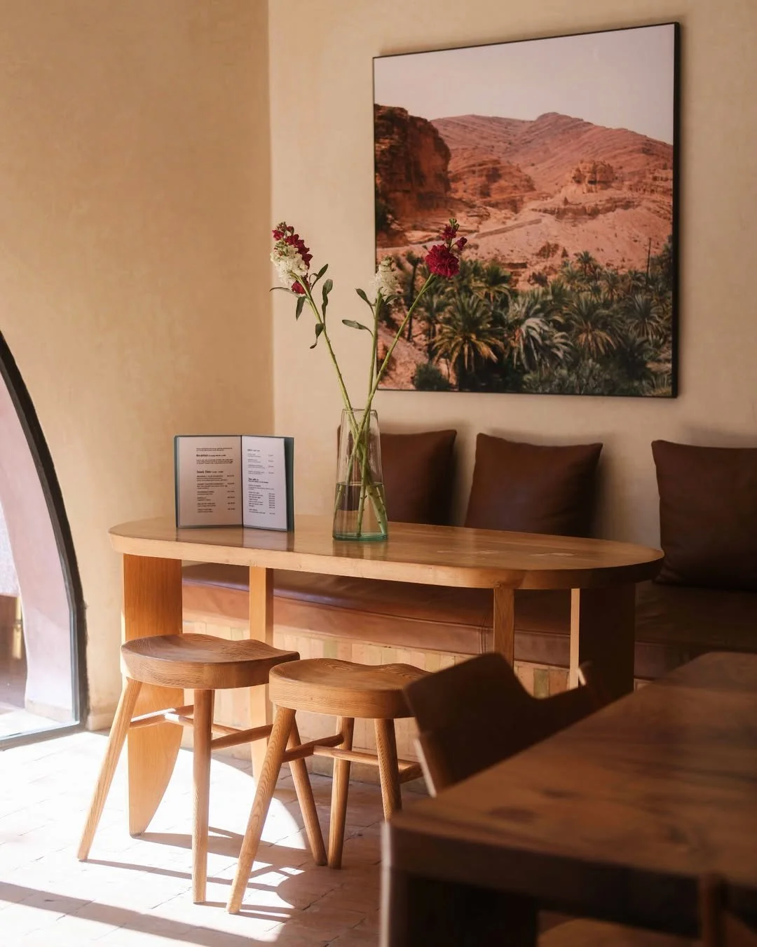 A cozy restaurant corner with a wooden table, two matching stools, and a vase with red and white flowers. A menu stand is on the table, and a leather bench with cushions is against the wall. A large framed desert landscape photograph hangs on the wall.
