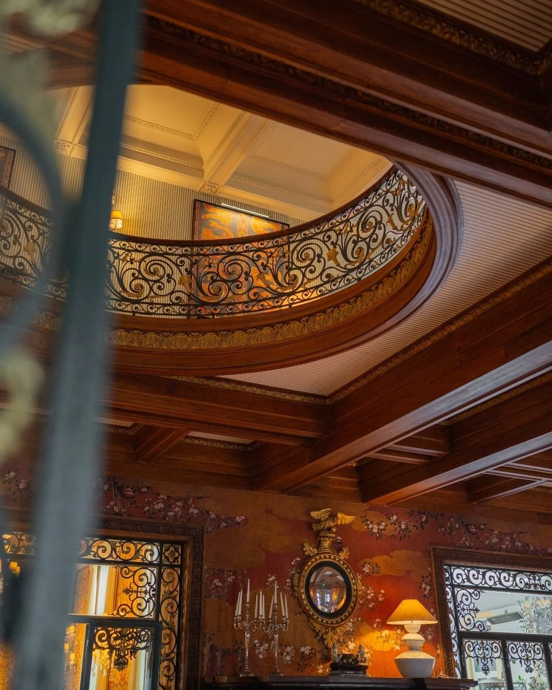 Interior view of a luxurious room with a wooden ceiling, decorative iron railings on the upper balcony, ornate mirror, floral wallpaper, chandelier, table lamp, and floral artwork.