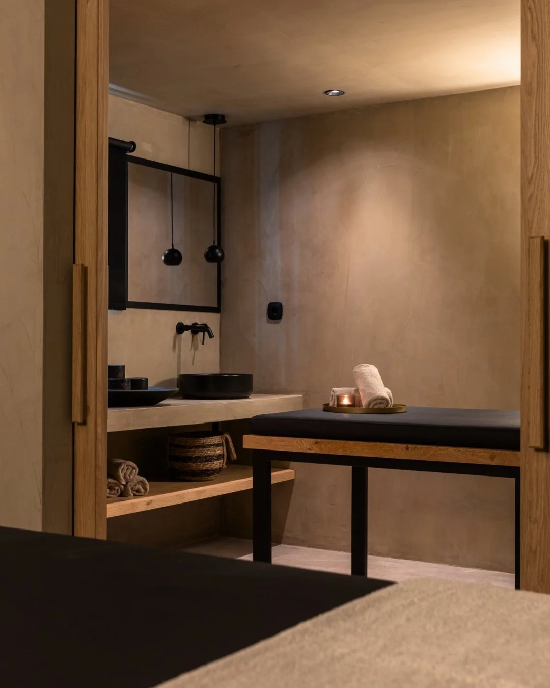 A minimalist spa treatment room with a massage table, rolled towels, a small candle, and a modern sink area with black accents.