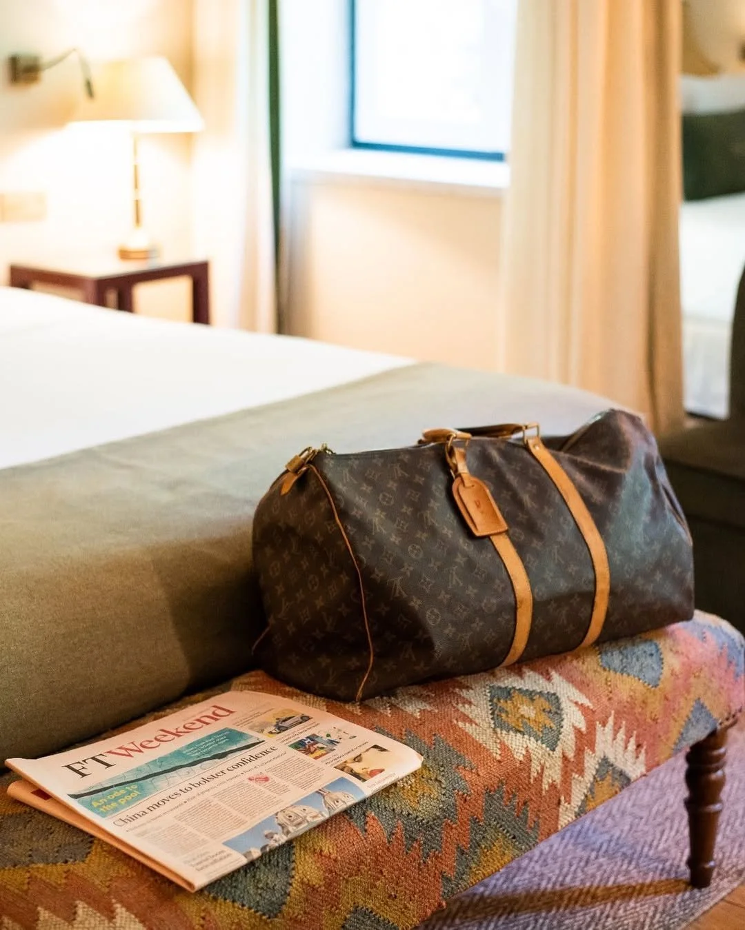 A hotel room with a bed, a bedside table with a lamp, a window with curtains, a patterned bench, a Louis Vuitton duffel bag, and a newspaper on the bench.