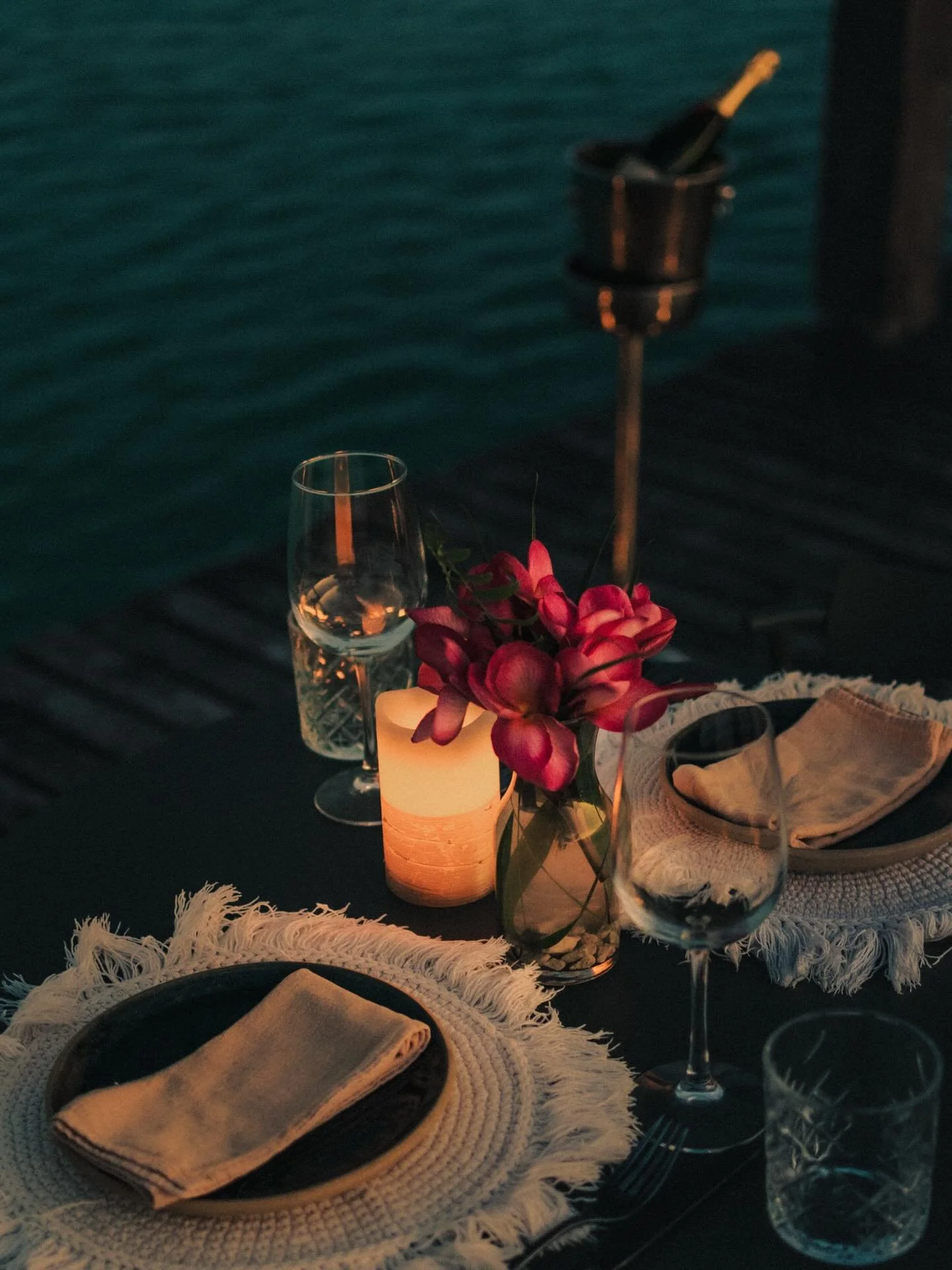 A table set for dinner with a white candle, a vase of pink flowers, a wine glass, and a plate with a beige napkin on top, against a dark water background.