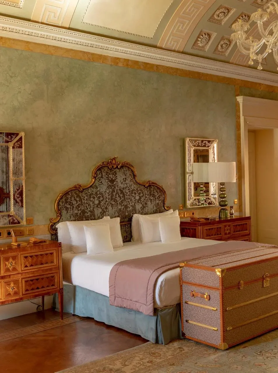 Luxurious hotel bedroom with ornate headboard, white bedding, decorative wall accents, and vintage-style furniture and decor.