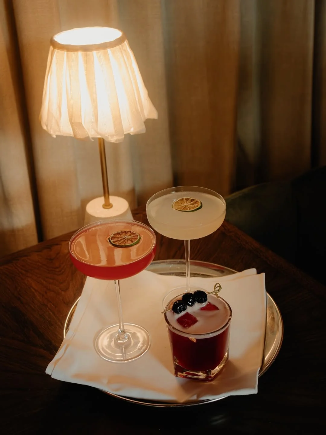 Three cocktails on a tray with a white napkin, positioned on a dark wooden table beside a small lamp with a fabric shade, in a cozy indoor setting.