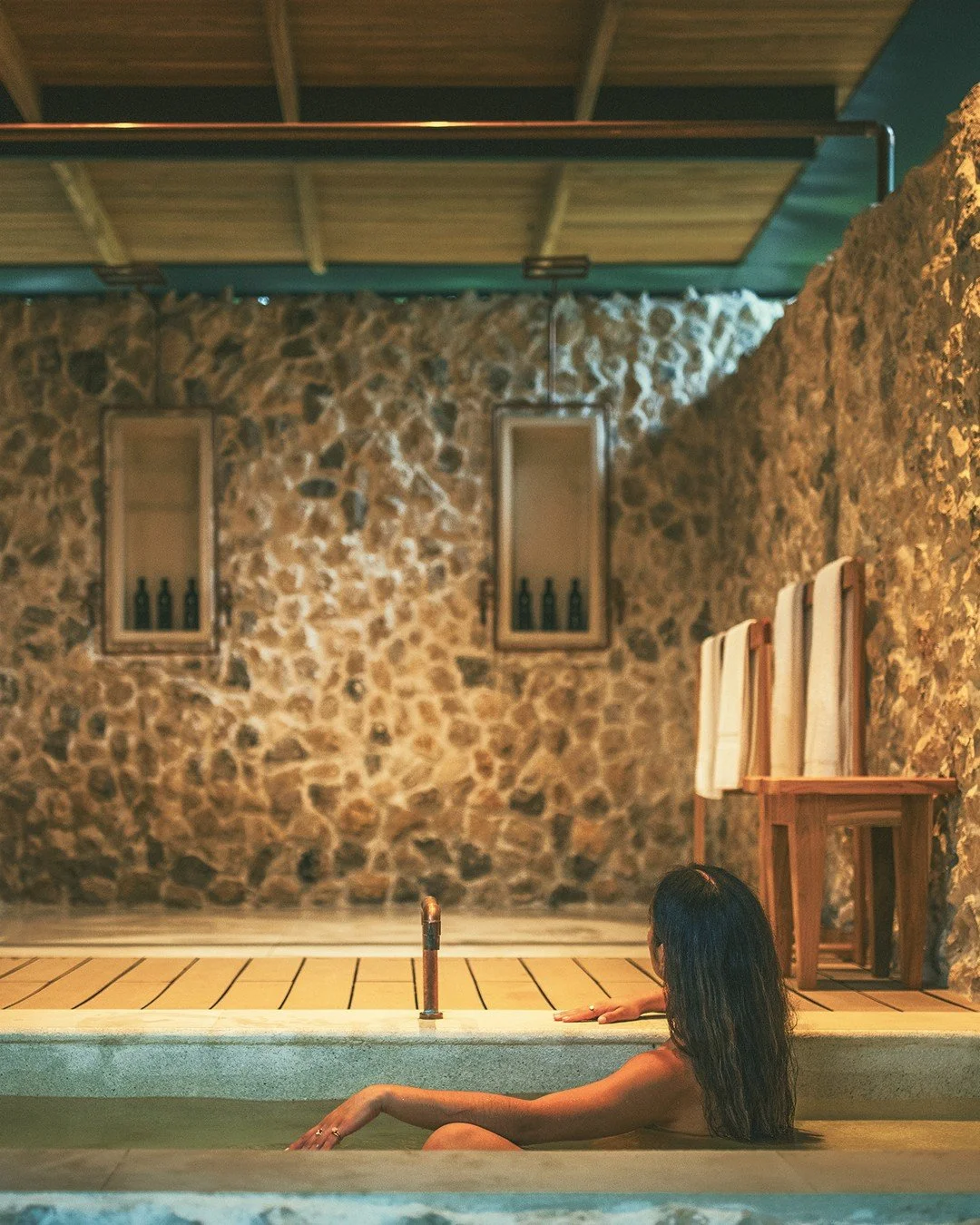 A woman relaxing in an indoor hot tub or spa with stone walls and wooden accents, facing away from the camera.