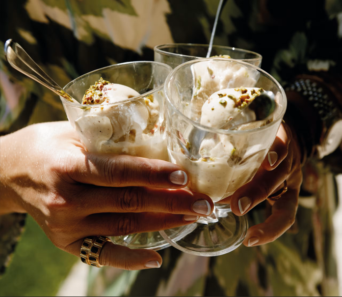 Three people holding glasses of ice cream with pistachios on top, clinking glasses in celebration.
