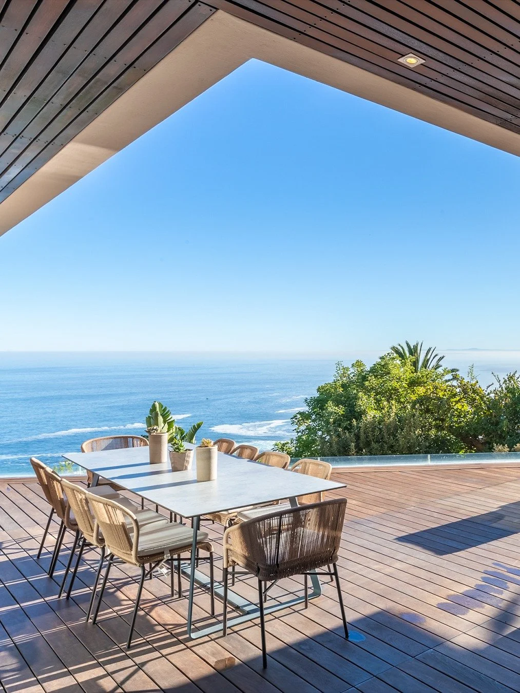 Ocean view from a balcony with a modern dining table and chairs, wooden flooring, and lush greenery.