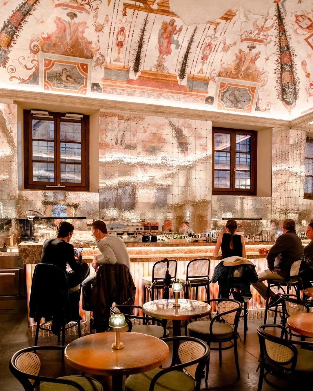 Interior of a stylish restaurant or bar with a high, ornate ceiling featuring classical artwork and frescoes. There are two windows on the back wall, a bar counter with people sitting and drinks, and small round tables with chairs in the foreground illuminated by small lamps.