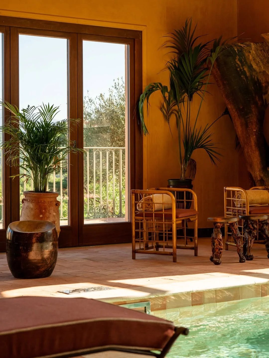 Indoor pool area with potted plants, wooden chairs, and a large window with a balcony view.