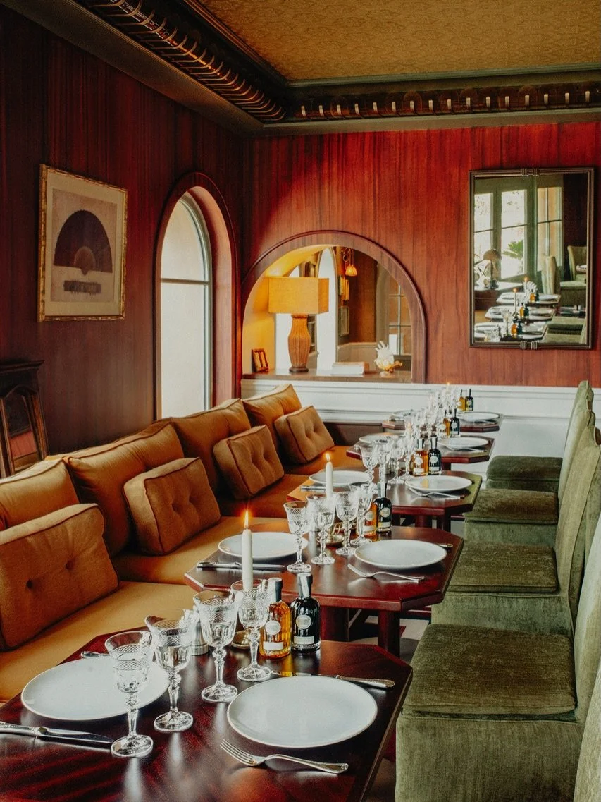 A cozy, elegant restaurant with a long dining table set with white plates, crystal glasses, candlelight, and small bottles, surrounded by plush green and tan upholstered chairs and red wooden paneled walls.