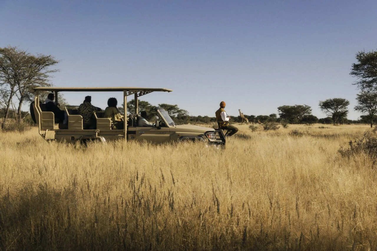 Tourists on an open-air safari vehicle viewing animals, including giraffes, in a savanna with tall grass and scattered trees under a clear blue sky.