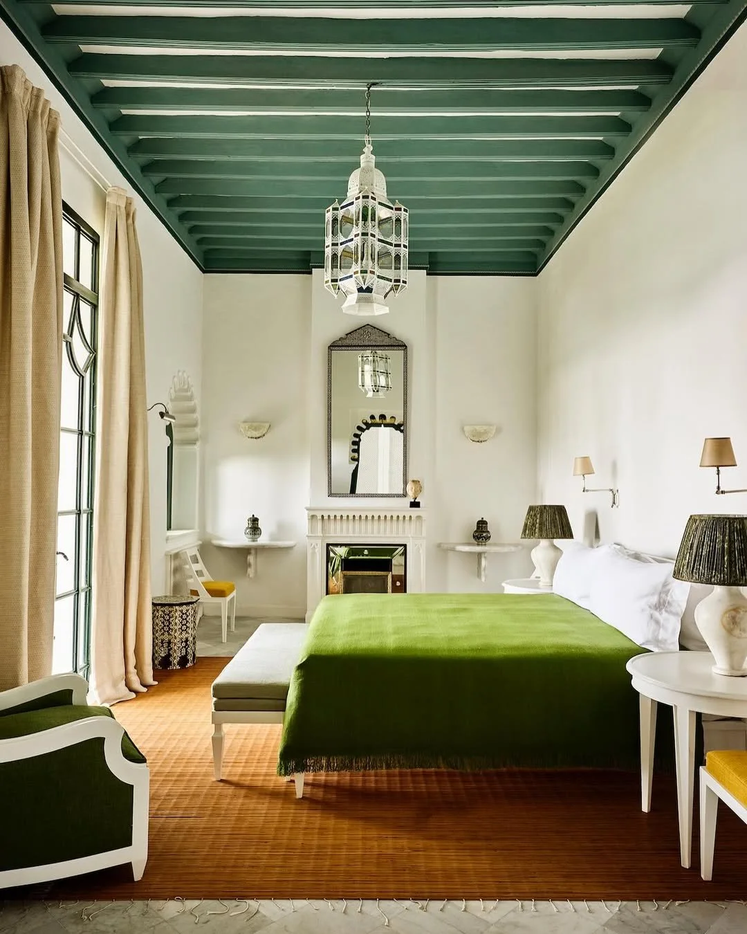 A bedroom with a high, teal ceiling, a green bedspread, and white walls. There are nightstands with lamps, a mirror above the fireplace, and beige curtains on large windows.