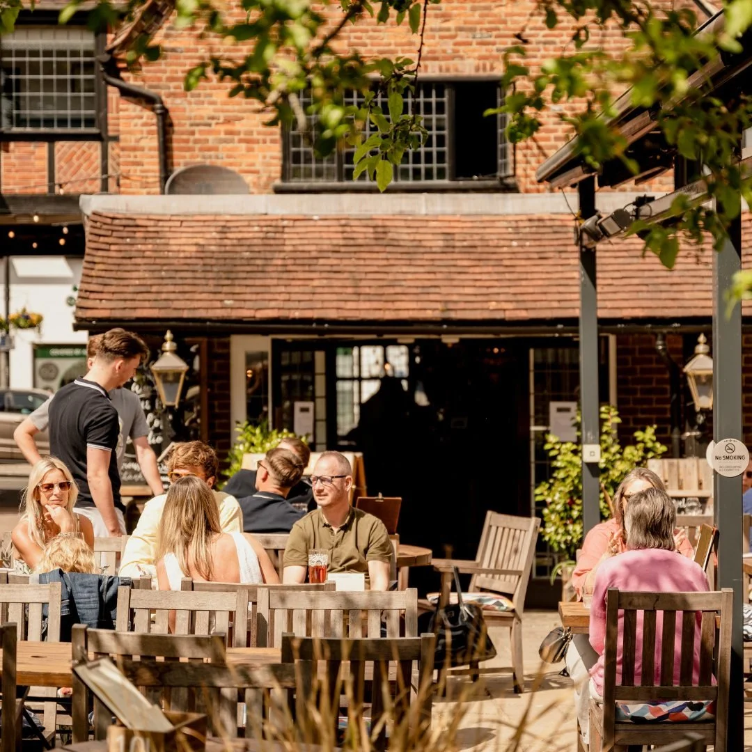 People dining outdoors at a restaurant patio with wooden tables and chairs, brick building in the background, and trees overhead on a sunny day.