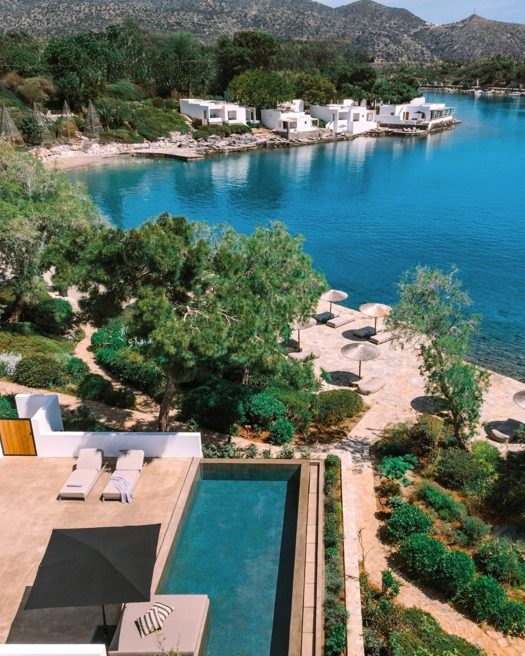 Aerial view of a luxurious lakeside property with a swimming pool, surrounded by lush greenery, modern white houses on the water, and a scenic mountain backdrop.
