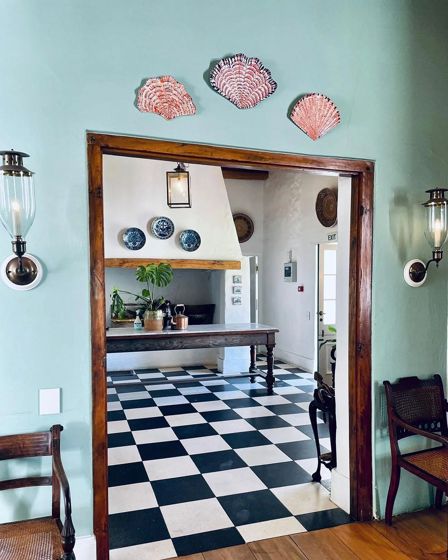 View of a dining area with a black and white checkered floor, green walls, wooden door frame, and wall-mounted lamps. Inside, there is a dining table with a plant and decorative items, and a wall with blue and white plates.