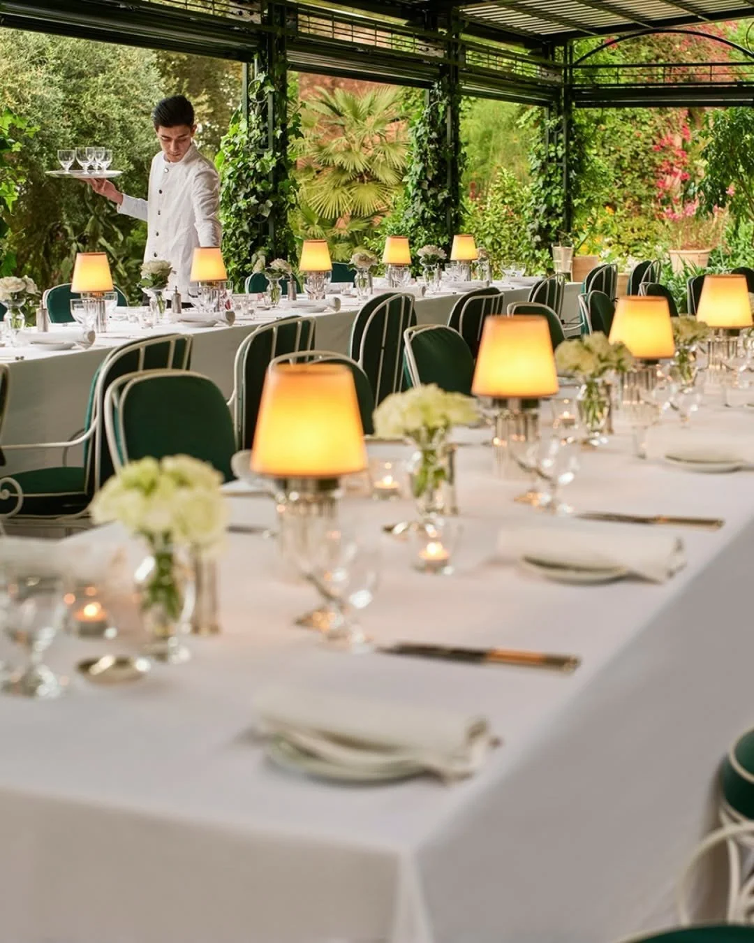 Elegant outdoor dining setup with long white tablecloths, small lamps, floral centerpieces, and place settings, surrounded by greenery and plants.
