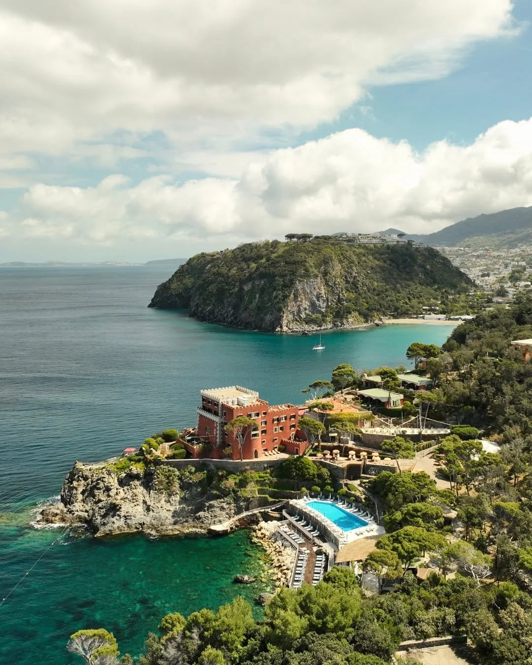 A coastal landscape with a large green hill extending into the ocean, houses on the hillside, and a swimming pool near the shoreline surrounded by trees, with a sailboat anchored nearby.