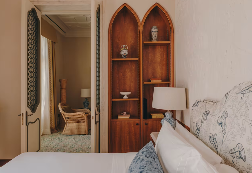 A bedroom with a built-in wooden bookshelf, a bed with patterned pillows, and a nightstand with a table lamp. Another room with a wicker chair, a lamp, and tall decorative vases is visible through an open door.