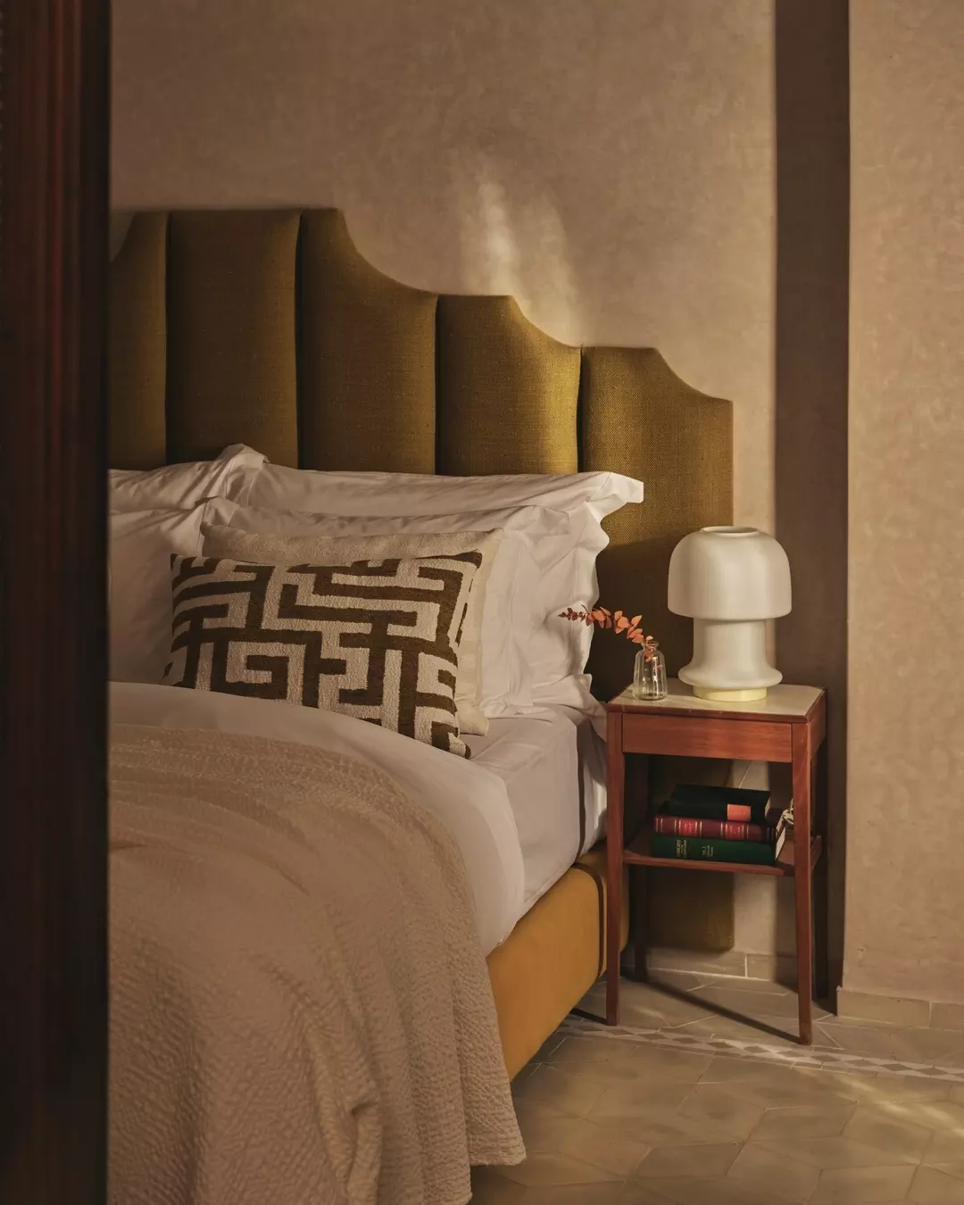 A cozy bedroom corner with a bed, decorative pillow, nightstand, lamp, and a small vase with a branch, in warm lighting.