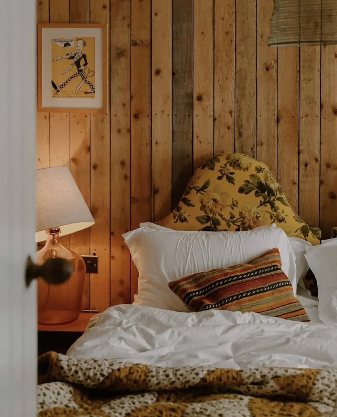A cozy bedroom with wooden paneled walls, a floral headboard, white bedding, a striped pillow, a bedside lamp, and framed artwork.