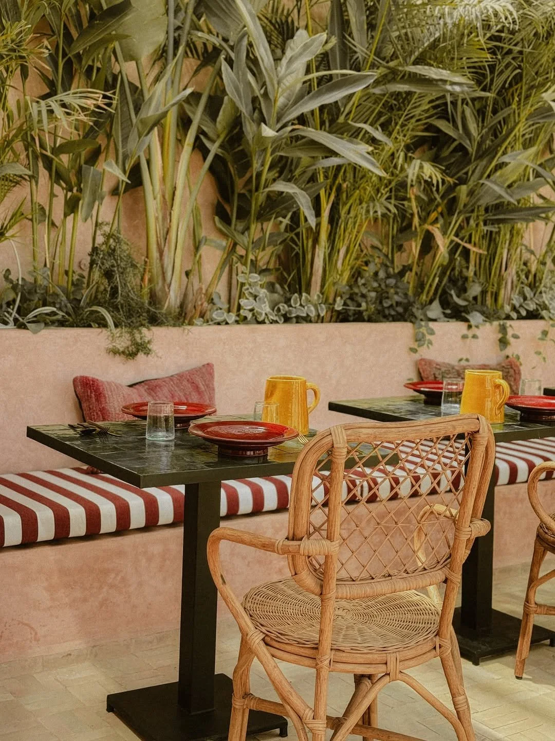 A restaurant table set with red plates, yellow pitchers, clear glasses, and silverware, with striped bench seating, rattan chairs, and a lush green plant wall in the background.