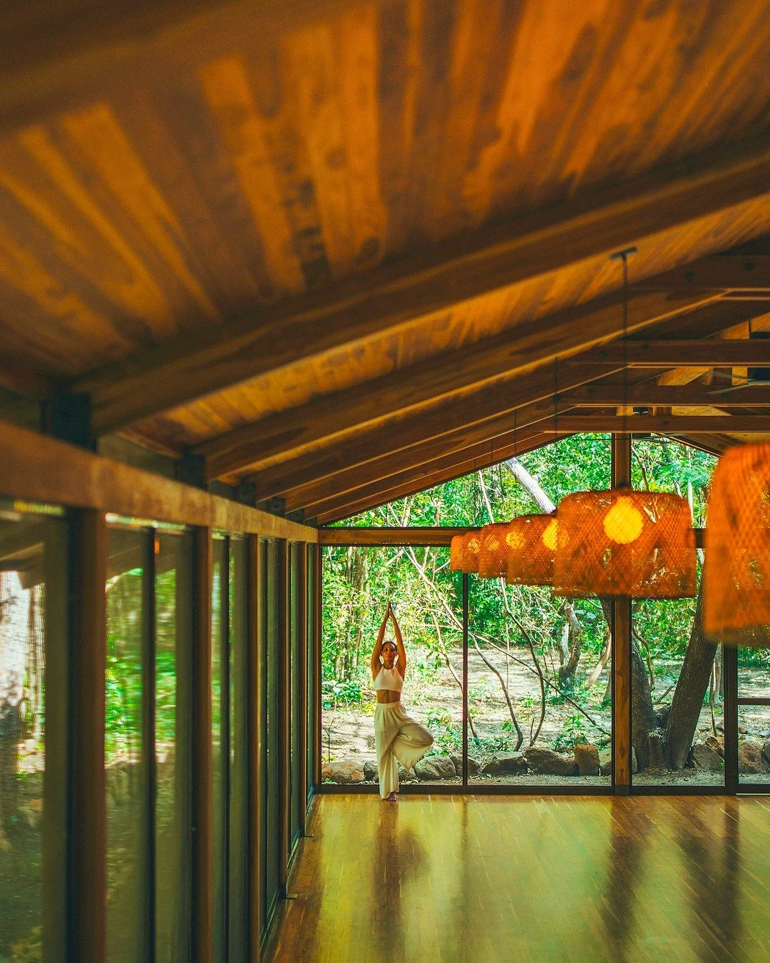 A woman practicing yoga in a spacious room with wooden ceiling and floors, large glass windows showing greenery outside, and hanging pendant lights.