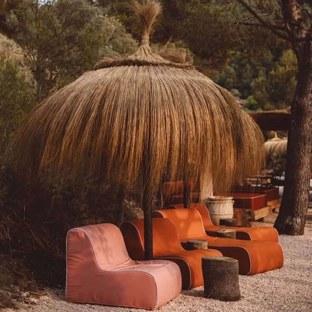 Outdoor seating area with pink and orange chairs under a large thatched umbrella, surrounded by trees.