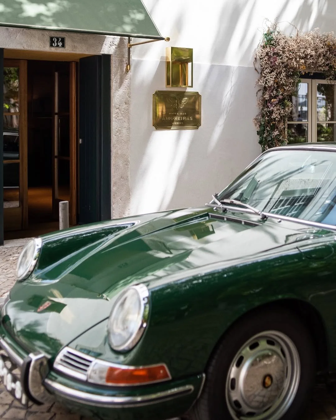 A vintage green Porsche car parked in front of a building with a white exterior wall, flowers, and a gold sign that reads 'Hotel das Amoreiras Lisbon'.
